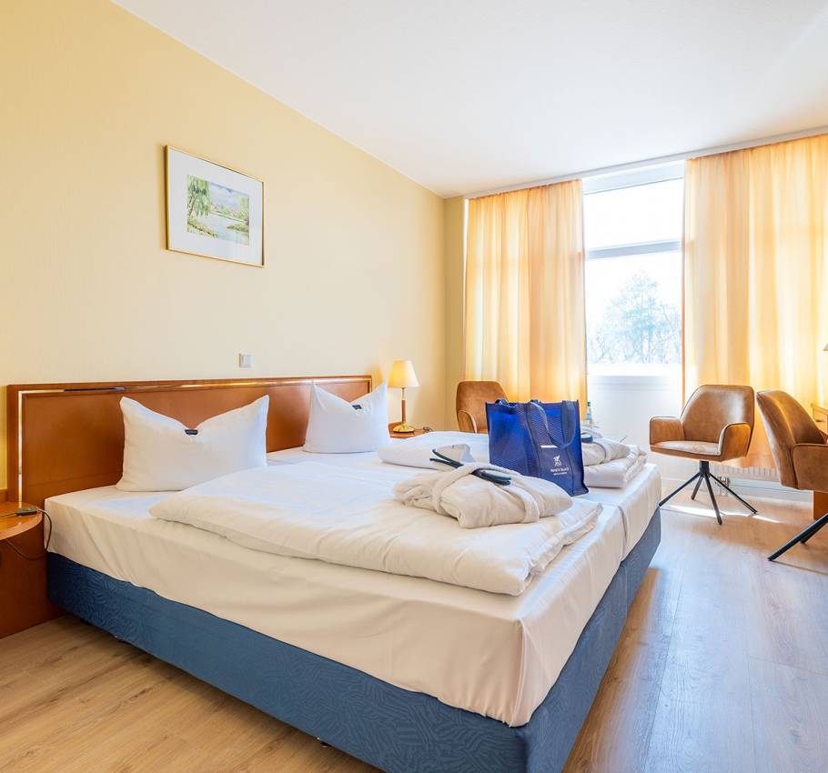 Doppelzimmer im Arkona Strandhotel Binz mit Blick auf das Bett, eine Sitzecke und ein großes Fenster mit Stadtblick.