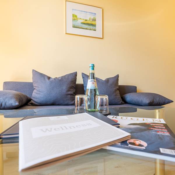 Sofa with decorative cushions, glass table with a water bottle and magazines in the suite of Arkona Strandhotel Binz, perfect for relaxation.