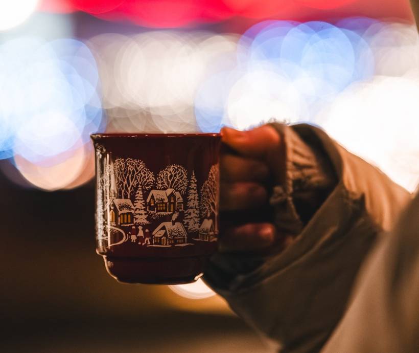 Person hält eine weihnachtlich verzierte Tasse mit Heißgetränk vor stimmungsvollen Lichtern und festlicher Weihnachtsbeleuchtung.