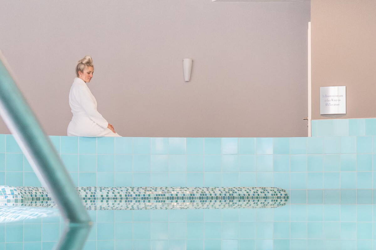 Eine blonde Frau sitzt am Beckenrand des Hotelpools und beobachtet das Wasser.