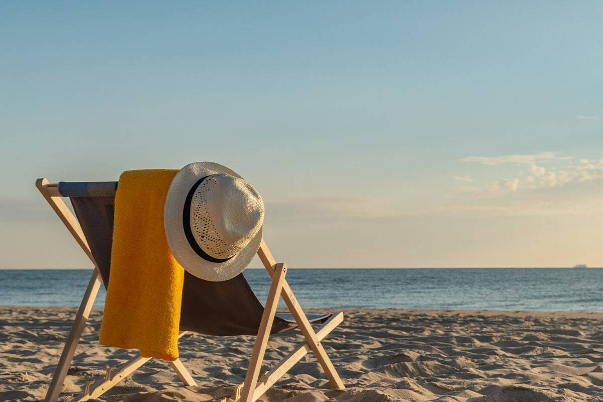 In der Sonne steht ein Klappstuhl am Strand, darüber hängt ein gelbes Handtuch und ein Hut.