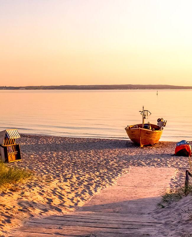 Ein hölzernes Boot liegt im Sonnenuntergang am Strand, daneben zwei Strandkörbe.
