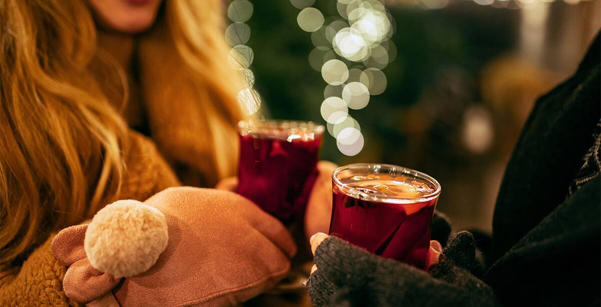 Zwei Frauen stehen lächelnd auf dem Weihnachtsmarkt und stoßen mit Glühwein an.