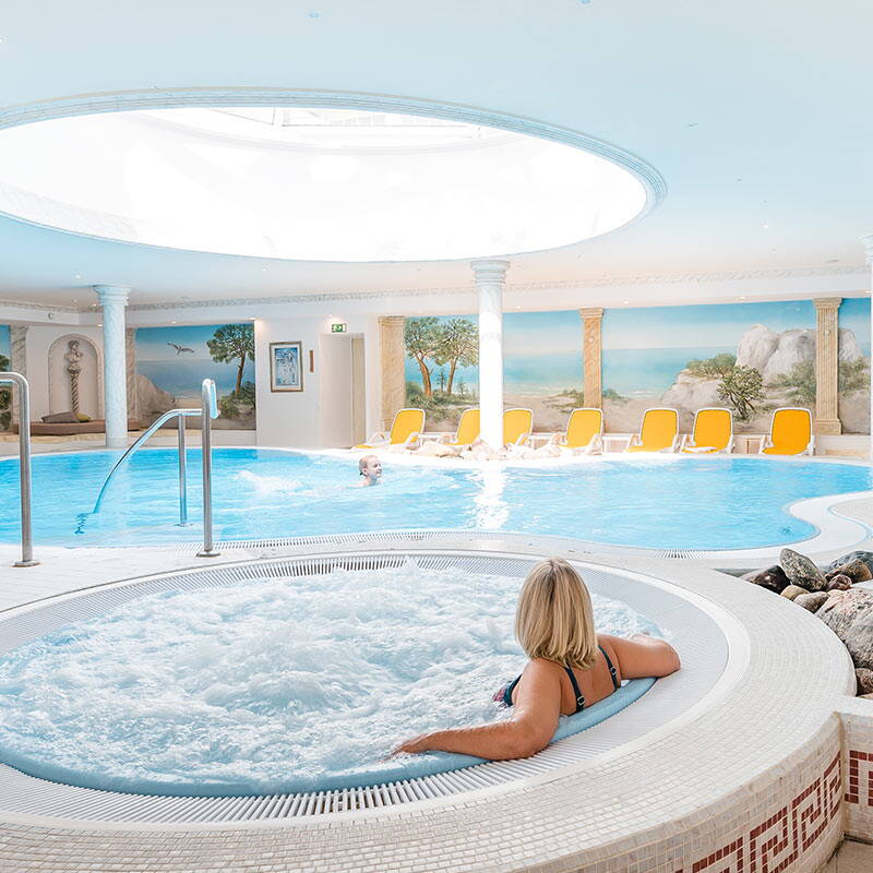Eine Frau sitzt entspannt im Whirlpool im Wellnessbereich des Hotels, im Hintergrund ein großer Pool.