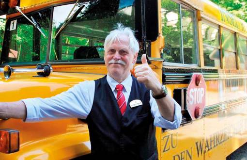 Ein Mann in Uniform lächelt in die Kamera und steht vor einem gelben Schulbus bereit für einen Ausflug des Arkona Strandhotels.