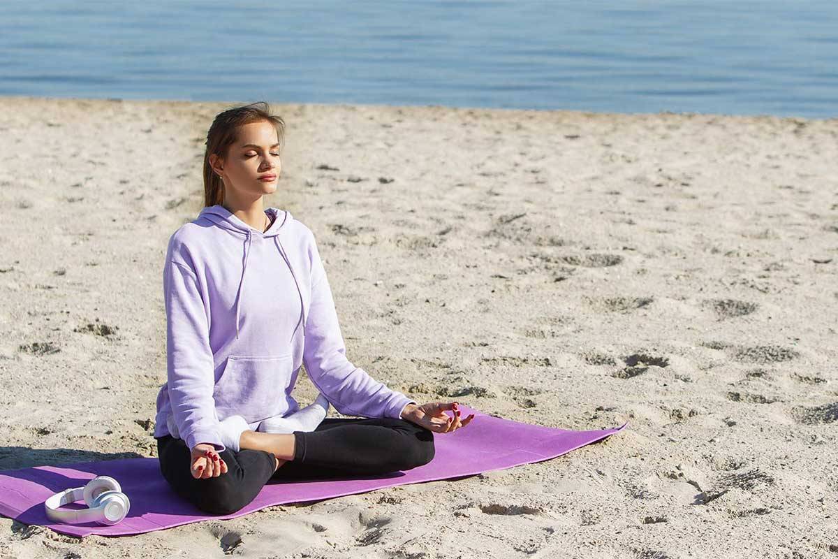 Eine junge Frau in violetten Sportklamotten sitzt am Strand und macht Yoga im Ostseeurlaub im Arkona Strandhotel.