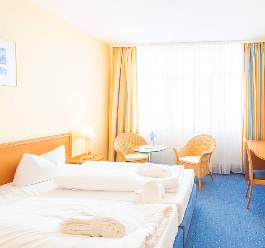 Doppelzimmer im Erdgeschoss des Arkona Strandhotels mit Blick auf das Bett, Sitzgelegenheit und Fenster mit Vorhängen.
