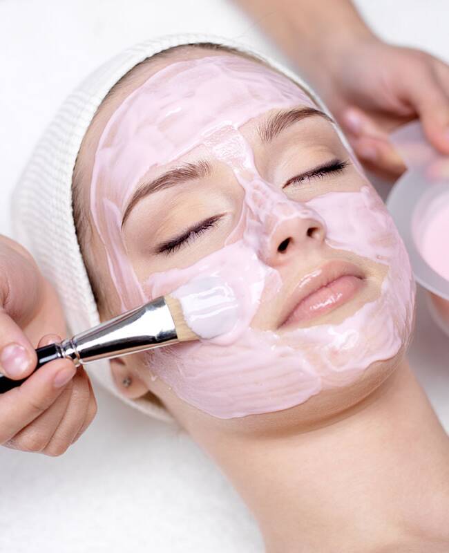 Eine rosa Maske wird mit einem Pinsel auf das Gesicht einer jungen Frau aufgetragen.