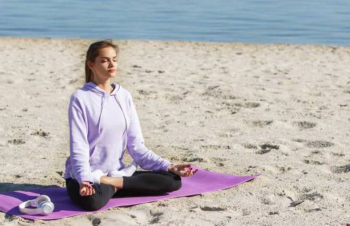 Eine junge Frau in violetten Sportklamotten sitzt am Strand und macht Yoga im Ostseeurlaub im Arkona Strandhotel.