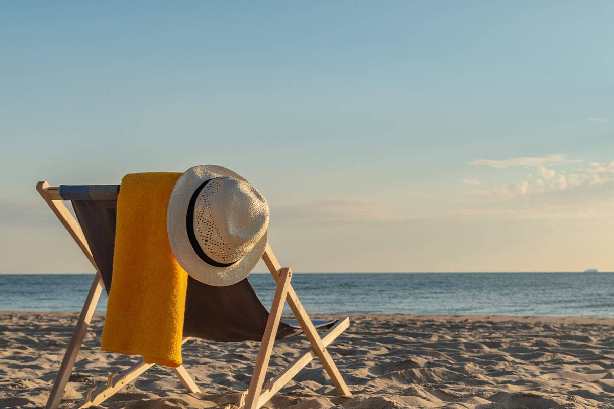 In der Sonne steht ein Klappstuhl am Strand, darüber hängt ein gelbes Handtuch und ein Hut.