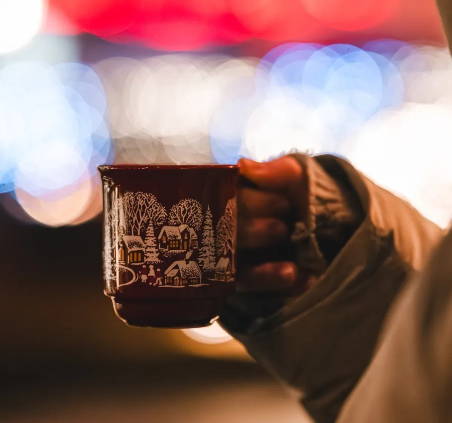 Person holding a festive Christmas mug filled with a hot drink against a background of warm holiday lights and seasonal decorations.