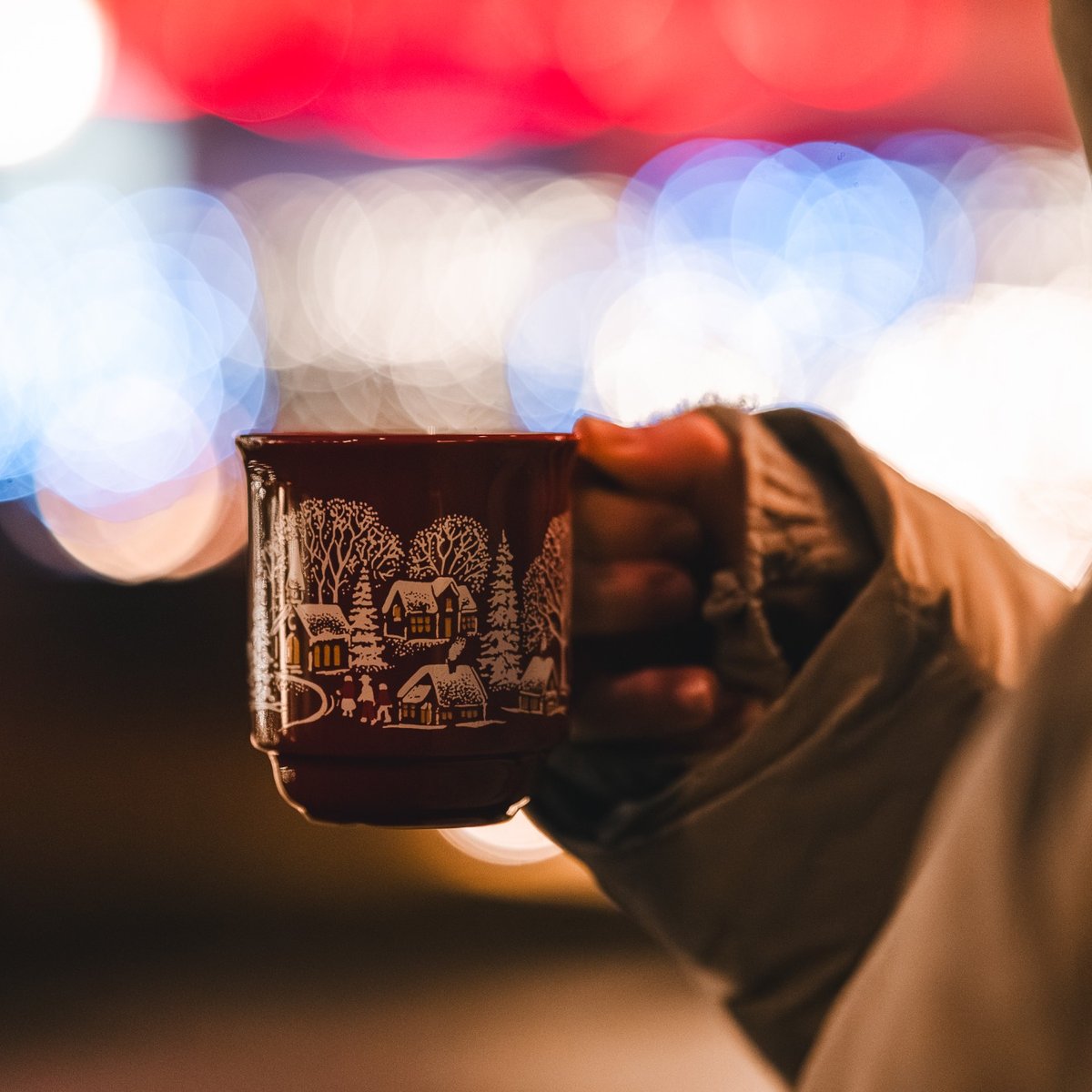 Deutsch   Weihnachtliche Tasse mit Heißgetränk in festlicher Atmosphäre (Titel)  Person hält eine weihnachtlich verzierte Tasse mit Heißgetränk vor stimmungsvollen Lichtern und festlicher Weihnachtsbeleuchtung.
