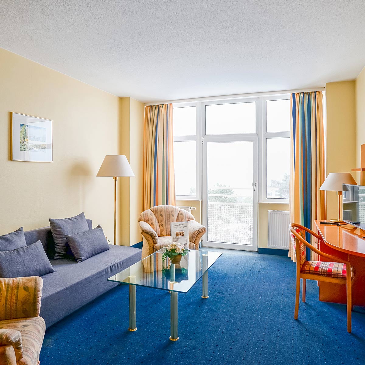 Wohnbereich mit grauem Sofa, Sessel, Glas-Couchtisch, Schreibtisch und großem Fenster.