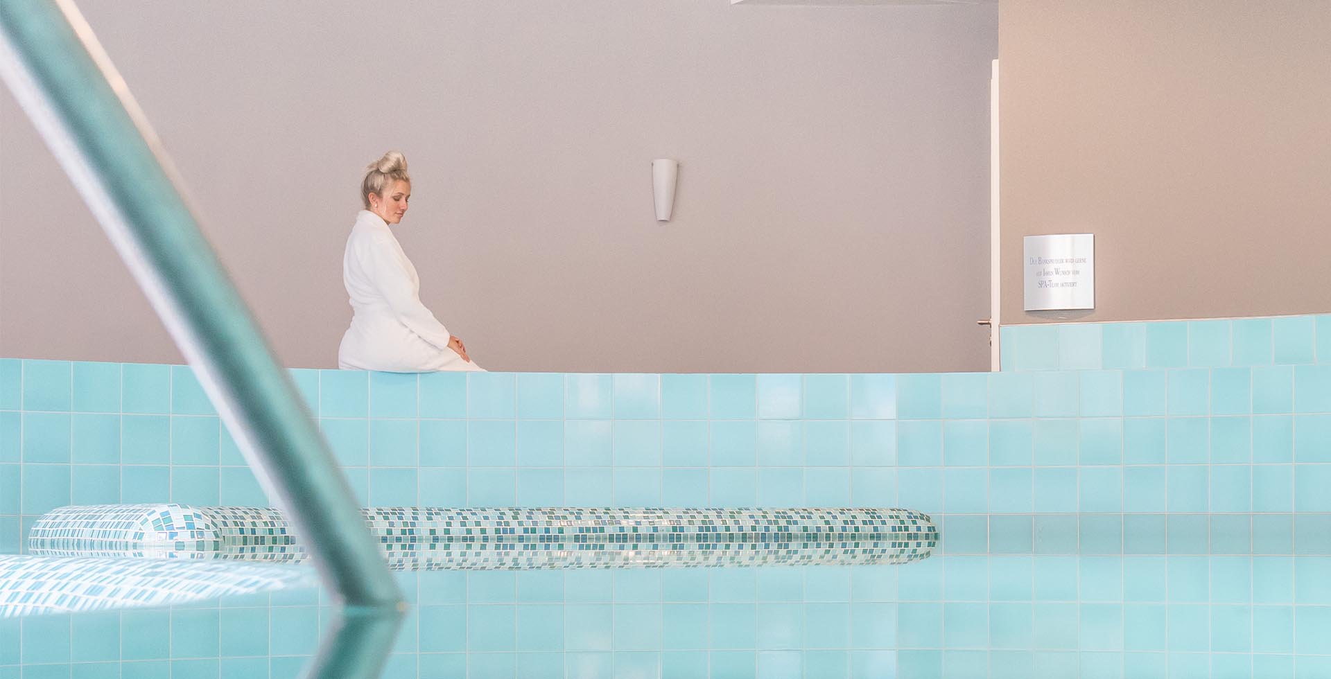 Eine blonde Frau sitzt am Beckenrand des Hotelpools und beobachtet das Wasser.
