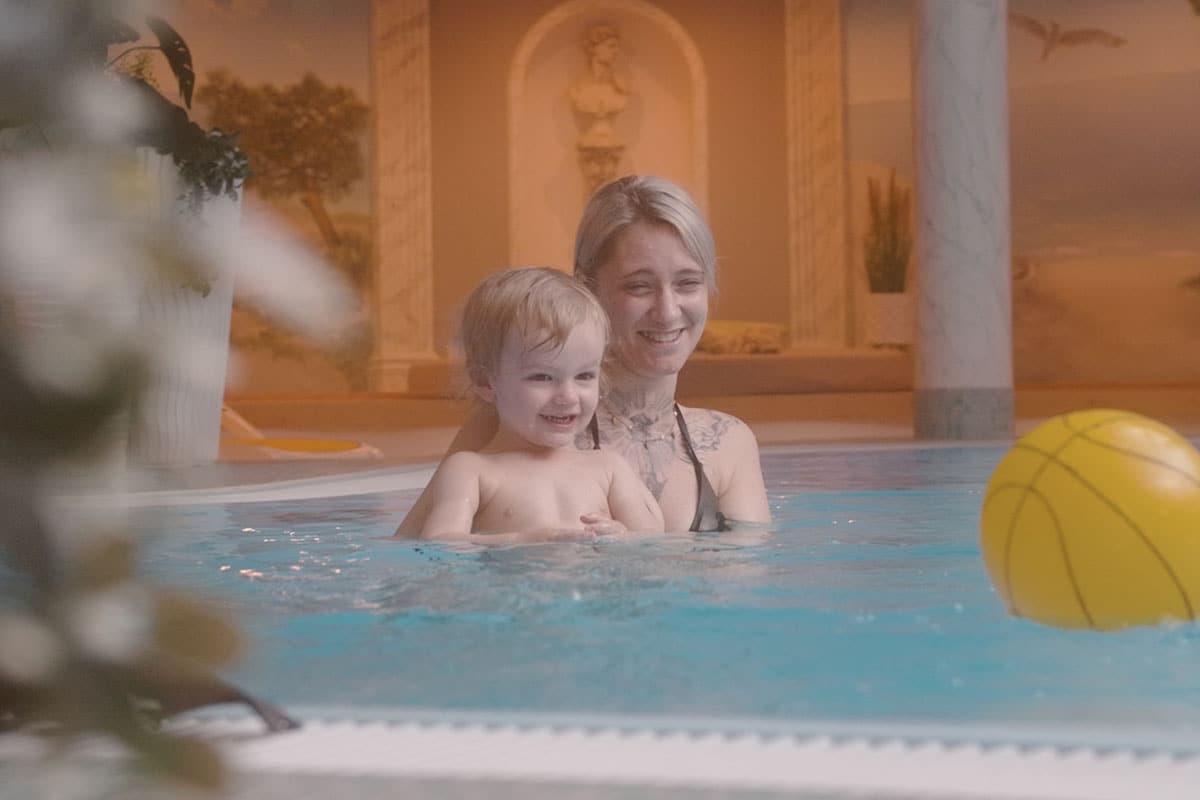 Eine Mutter entspannt sich im Whirlpool im Wellnessbereich während sie ihrer Tochter beim Schwimmen im Indoorpool zuschaut.