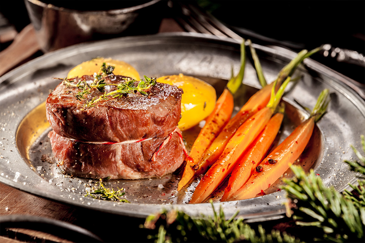 Gebratenes Filetsteak auf einem Metallteller, serviert mit glasierten Karotten und Kartoffeln.